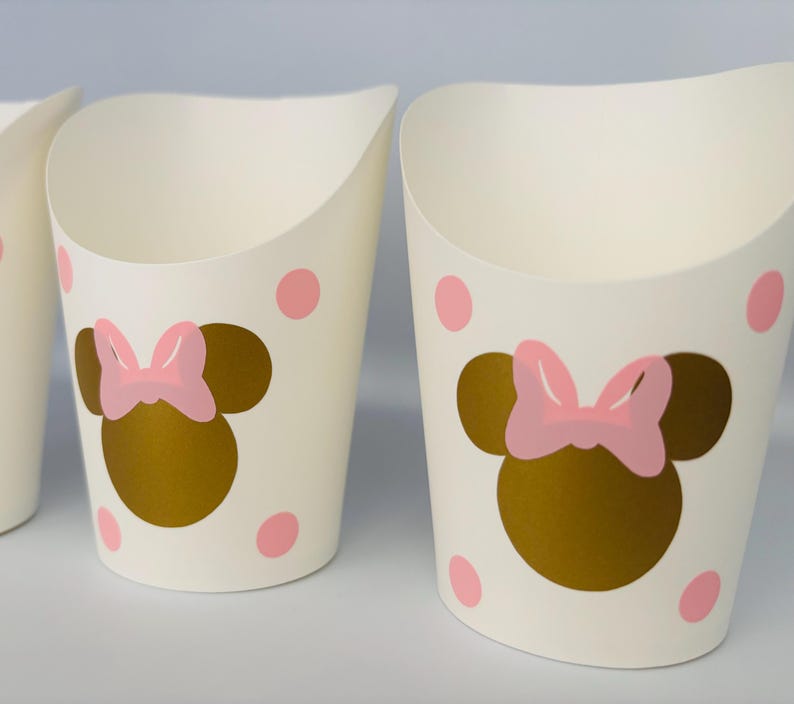 K&ouml;nnte beinhalten: Drei wei&szlig;e Pappbecher mit einer goldenen Minnie Mouse-Silhouette und einer rosa Schleife. Die Becher haben rosa Punkte.