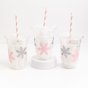 May include: Three clear plastic cups with pink and gray snowflake designs. Each cup has a pink and white striped paper straw.