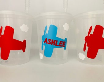 May include: Three clear plastic cups with straws, each decorated with a different airplane design. One cup has a red airplane, one has a blue airplane with the name "ASHLEE" in red letters, and the other has a red airplane.