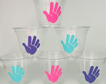 May include: Six clear plastic cups with handprint designs. The handprints are in pink, blue, and purple.