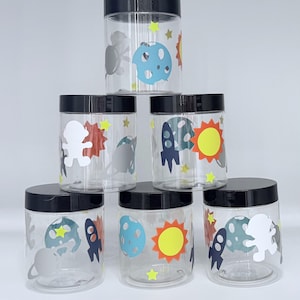 May include: A set of six clear plastic cylindrical storage jars with black screw-on lids. Each jar features colorful vinyl decals of space-themed elements including yellow stars, orange suns, blue planets, gray Saturn-like rings, and dark blue rockets.