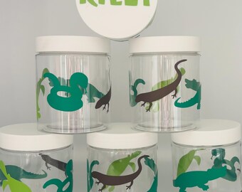 May include: Clear plastic jars with white lids, decorated with green and brown lizard and reptile designs. The lid of one jar has the name "RILEY" in green letters.