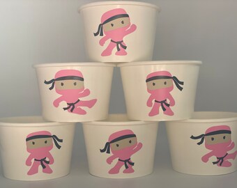 May include: Six white paper cups with pink ninja characters on them. The ninjas are wearing black belts and have black eyes. The cups are stacked in a pyramid shape.