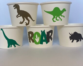 May include: Set of five white paper cups with dinosaur designs. The cups feature a green dinosaur, a brown dinosaur, a green dinosaur with the word "ROAR" in green, brown, and black letters, and a black dinosaur.