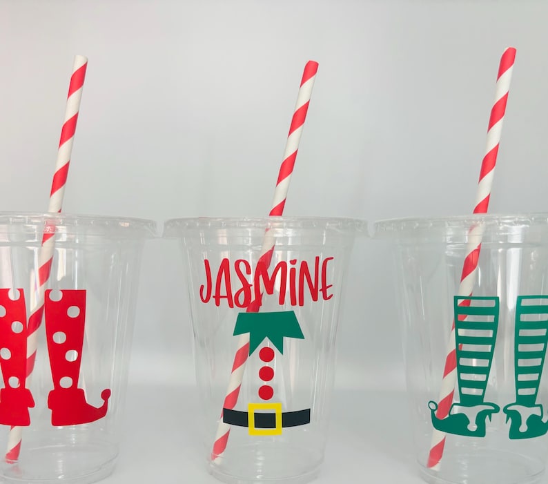 May include: Three clear plastic cups with red and white striped straws. Each cup has a different design featuring a Christmas elf. One cup has the name "Jasmine" and a green elf with a red hat and a yellow belt. Another cup has a red elf with a white hat and white boots. The third cup has a green elf with a white hat and white striped boots.