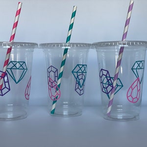 May include: Three clear plastic cups with lids and straws. Each cup has a different colored straw and a design of purple and pink diamonds on the side.