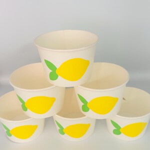 May include: Six white paper cups with a yellow lemon design. The lemons have green leaves. The cups are stacked in a pyramid shape.