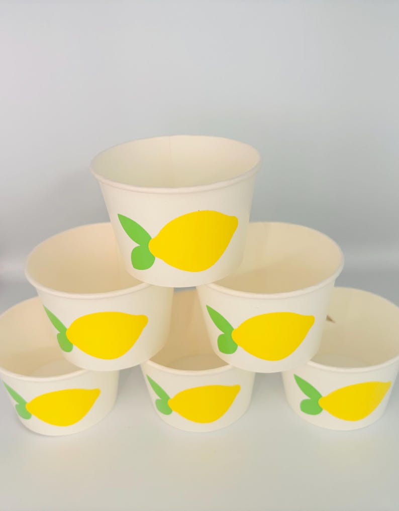 May include: Six white paper cups with a yellow lemon design. The lemons have green leaves. The cups are stacked in a pyramid shape.