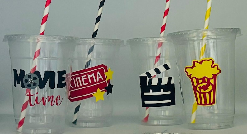 May include: Four clear plastic cups with red and black striped straws. Each cup has a different movie-themed design: "Movie time", "Cinema", a film clapboard, and a bucket of popcorn.