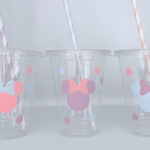 May include: Three clear plastic cups with lids, each featuring a Minnie Mouse design in pink, purple, and blue. The cups have matching polka dots and striped straws. A fun and colorful party supply.