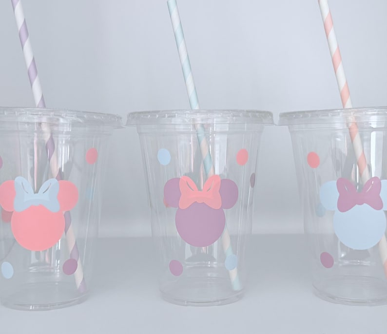 May include: Three clear plastic cups with lids, each featuring a Minnie Mouse design in pink, purple, and blue. The cups have matching polka dots and striped straws. A fun and colorful party supply.