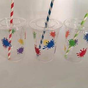 May include: Three clear plastic cups with colorful paint splatter designs and matching striped straws. The cups are perfect for a party or celebration.