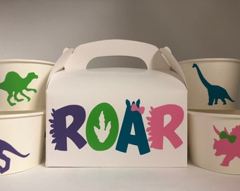 May include: A set of four white paper party favor cups with dinosaur silhouettes. The cups feature a green dinosaur, a blue dinosaur, a purple dinosaur, and a pink dinosaur. The cups are packaged in a white box with a handle and the word "ROAR" in colorful letters.