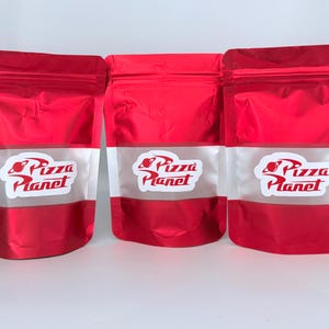 May include: Three red food pouches with a white band featuring the "Pizza Planet" logo. The logo includes a planet graphic and stylized text. The pouches are sealed and stand upright against a white background.