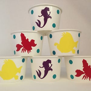 May include: Six white paper cups with teal polka dots and different coloured character silhouettes. The characters include a mermaid, a crab, and a fish.