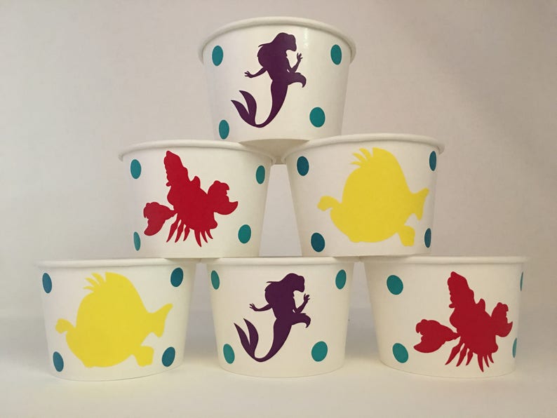 May include: Six white paper cups with teal polka dots and different coloured character silhouettes. The characters include a mermaid, a crab, and a fish.