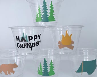 May include: Clear plastic cups with camping-themed designs. The cups feature illustrations of trees, a campfire, a bear, and a tent. One cup has the text "HAPPY camper" in black. These cups are perfect for a camping-themed party.