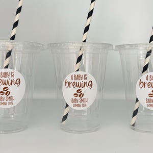 May include: Three clear plastic cups with lids, each featuring a black and white striped straw. Each cup has a white sticker with the text "A BABY IS brewing" and "BABY SMITH COMING 2026" in brown font, along with coffee bean graphics.