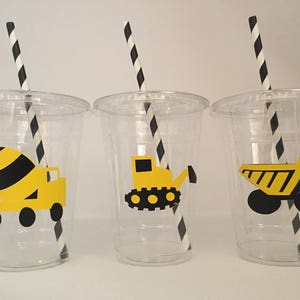 May include: Three clear plastic cups with black and white striped straws. Each cup has a different yellow construction vehicle design: a cement mixer, a bulldozer, and a dump truck.