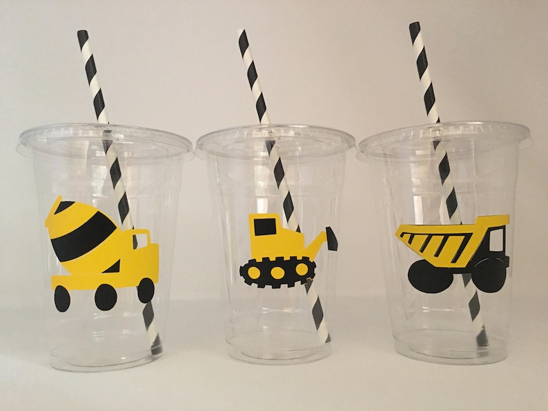 May include: Three clear plastic cups with black and white striped straws. Each cup has a different yellow construction vehicle design: a cement mixer, a bulldozer, and a dump truck.