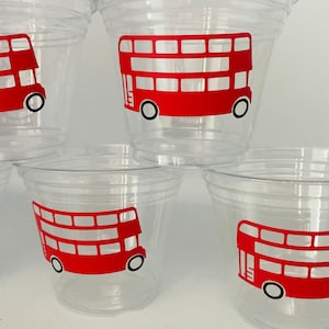 May include: Clear plastic cups with a red double-decker bus design. The cups are stacked and ready for use. The bus design features white windows and black wheels, perfect for a London-themed party or event.