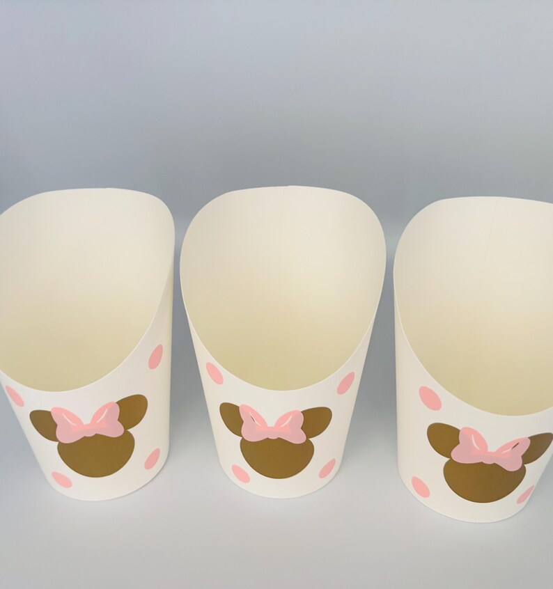 K&ouml;nnte beinhalten: Drei wei&szlig;e Pappbecher mit rosa Punkten und einem goldenen Minnie Mouse Kopf mit einer rosa Schleife auf jedem Becher.