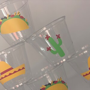 May include: Six clear plastic cups with colorful Mexican-themed designs. The designs include a yellow taco, a green cactus with red flowers, and a yellow sombrero.