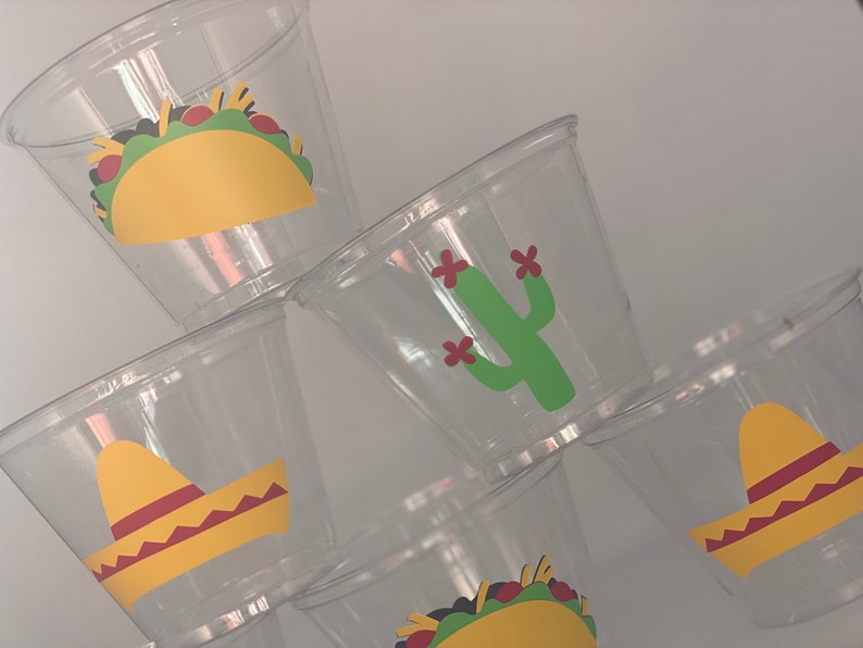 May include: Six clear plastic cups with colorful Mexican-themed designs. The designs include a yellow taco, a green cactus with red flowers, and a yellow sombrero.