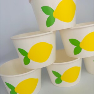 May include: A stack of six white paper cups with a yellow lemon and green leaf design. The cups are perfect for serving lemonade or other beverages at a summer party.