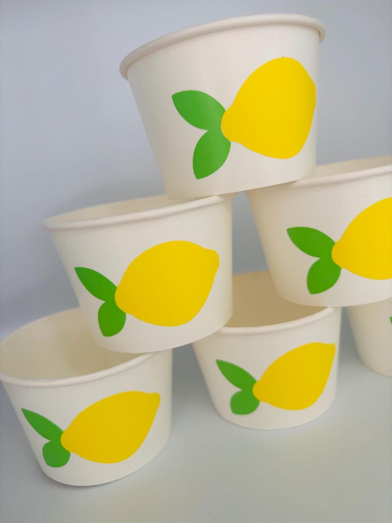 May include: A stack of six white paper cups with a yellow lemon and green leaf design. The cups are perfect for serving lemonade or other beverages at a summer party.