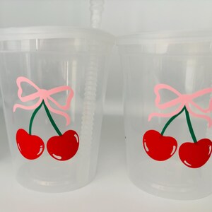 May include: Two clear plastic cups with lids and straws. Each cup has a pink bow and two red cherries with green stems printed on the side.