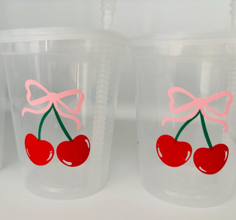 May include: Two clear plastic cups with lids and straws. Each cup has a pink bow and two red cherries with green stems printed on the side.