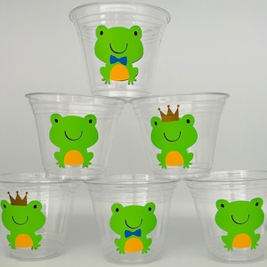 May include: Six clear plastic cups with frog designs. The frogs are green with yellow bellies and have different expressions. Some frogs wear a crown or a blue bow tie.