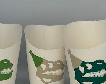 May include: Three white paper cups with dinosaur skull designs. Each cup has a green party hat on top of the skull. One cup has a green skull, one has a tan skull, and one has a green skull with a white outline.