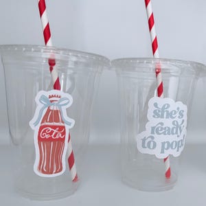May include: Two clear plastic cups with lids, each featuring a red and white striped straw. One cup has a sticker of a red Cola bottle with a blue bow, the other says "she's ready to pop!" in light blue.