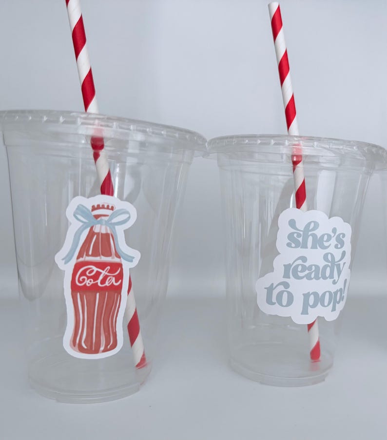 May include: Two clear plastic cups with lids, each featuring a red and white striped straw. One cup has a sticker of a red Cola bottle with a blue bow, the other says "she's ready to pop!" in light blue.