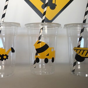 May include: Three clear plastic cups with black and white striped straws. Each cup has a yellow construction vehicle sticker on the front. The stickers are of a bulldozer, a cement mixer, and a dump truck.