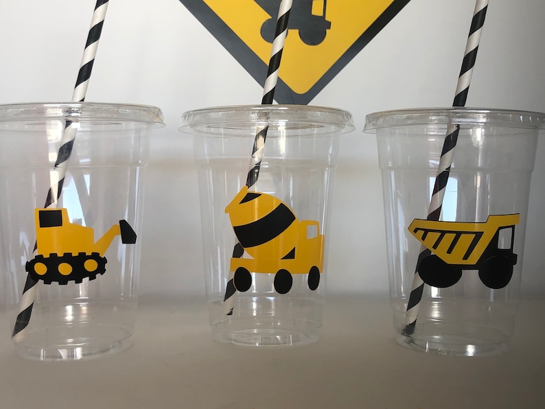 May include: Three clear plastic cups with black and white striped straws. Each cup has a yellow construction vehicle sticker on the front. The stickers are of a bulldozer, a cement mixer, and a dump truck.