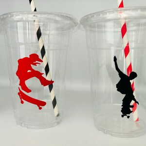 Puede incluir: Dos vasos de pl&aacute;stico transparentes con pajitas rayadas en blanco y negro. Un vaso tiene una silueta roja de un patinador, el otro tiene una silueta negra de un patinador.