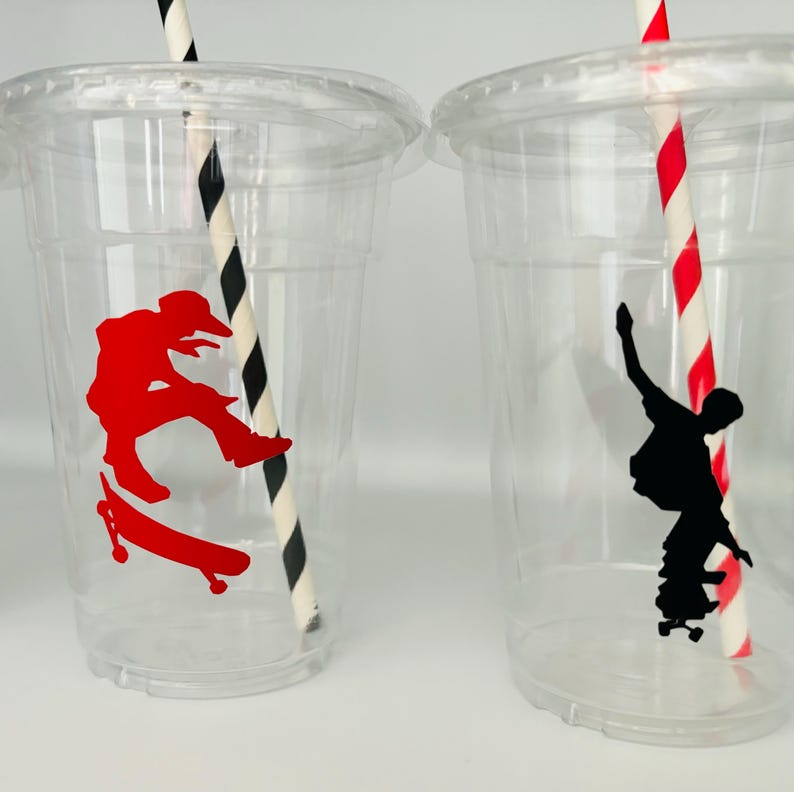 Puede incluir: Dos vasos de pl&aacute;stico transparentes con pajitas rayadas en blanco y negro. Un vaso tiene una silueta roja de un patinador, el otro tiene una silueta negra de un patinador.