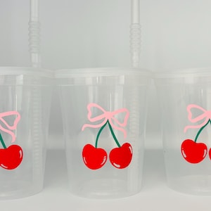 May include: Three clear plastic cups with straws and a pink bow and cherry design. The cherries are red with green stems and leaves.
