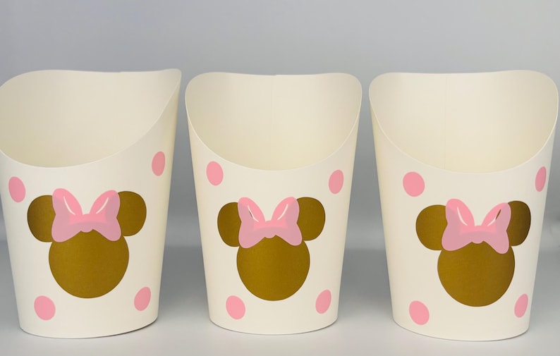 K&ouml;nnte beinhalten: Drei wei&szlig;e Pappbecher mit rosa Punkten und einem goldenen Minnie Mouse Kopf mit einer rosa Schleife auf jedem Becher.