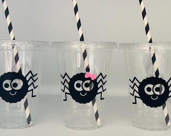 May include: Three clear plastic cups with black and white striped straws. Each cup has a black spider with googly eyes and a zigzag pattern around the edge. One spider has a pink bow.