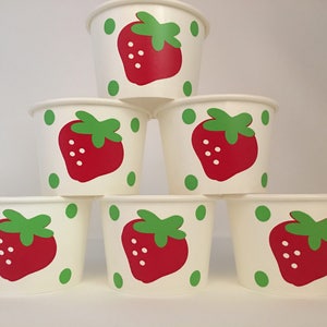 May include: Six white paper cups with a red strawberry and green leaves design. The cups are stacked in a pyramid shape.