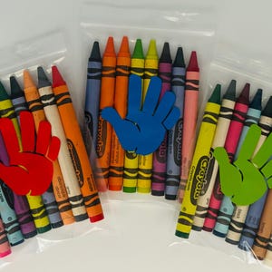 May include: Three clear plastic bags each containing a set of Crayola crayons. The bags are decorated with a handprint cutout in red, blue, and green.