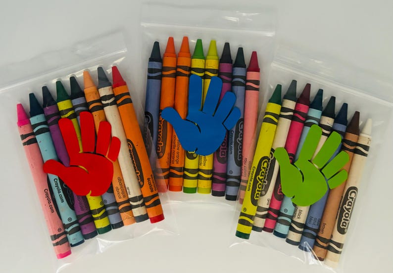 May include: Three clear plastic bags each containing a set of Crayola crayons. The bags are decorated with a handprint cutout in red, blue, and green.