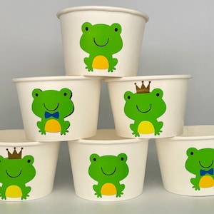 May include: Six white paper cups with frog designs. The frogs are green with yellow bellies and some have a blue bow tie or a gold crown.