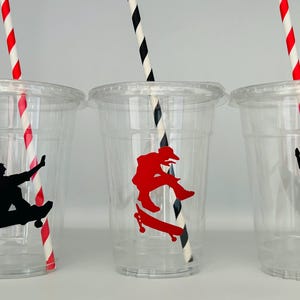 Puede incluir: Tres vasos de pl&aacute;stico transparentes con pajitas de rayas rojas y negras. Cada vaso tiene una silueta negra de una persona haciendo skate con una tabla de skate roja.