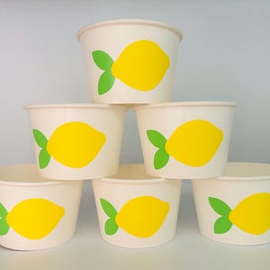 May include: Six white paper cups with yellow lemon designs. Each cup has a green leaf on the lemon.