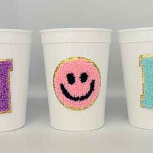 May include: Three white plastic cups with decorative patches. One cup has a purple letter "N" with gold trim, another has a pink smiley face with black eyes and mouth, and the last has a blue letter "B" with gold trim.